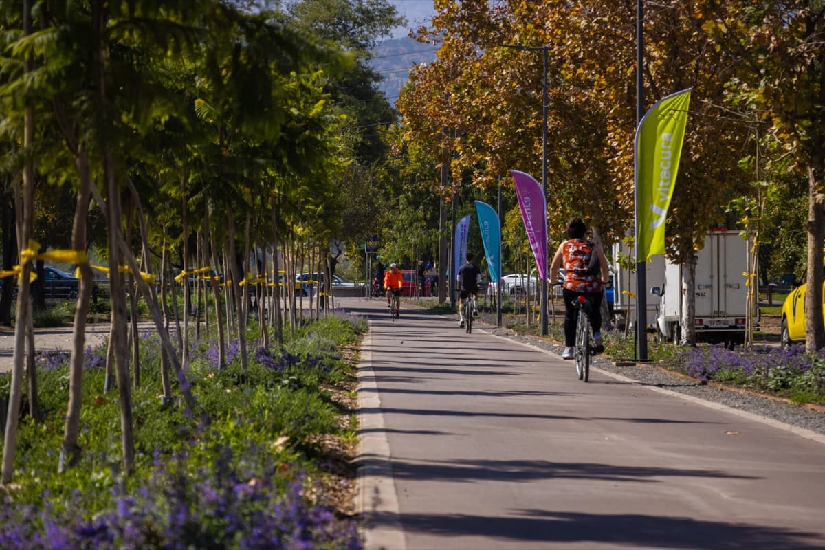 Ciclistas pedaleando por la ciclovía del río Mapocho en Santiago, con árboles y la cordillera de fondo